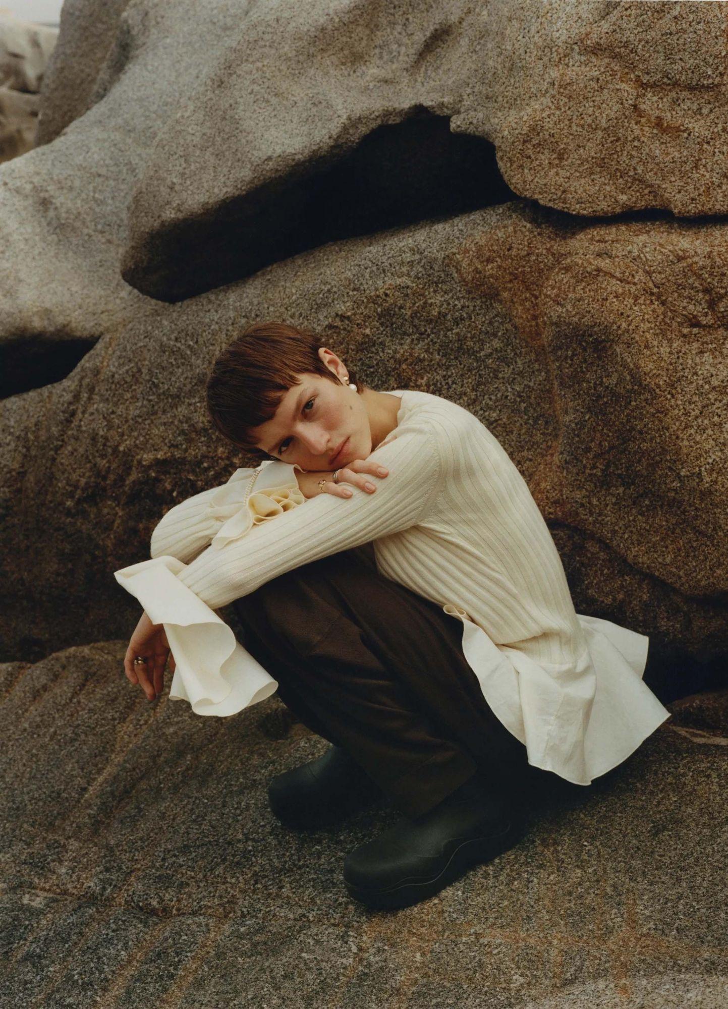 A model crouched against weathered granite in a cream ribbed knit with flared cuffs