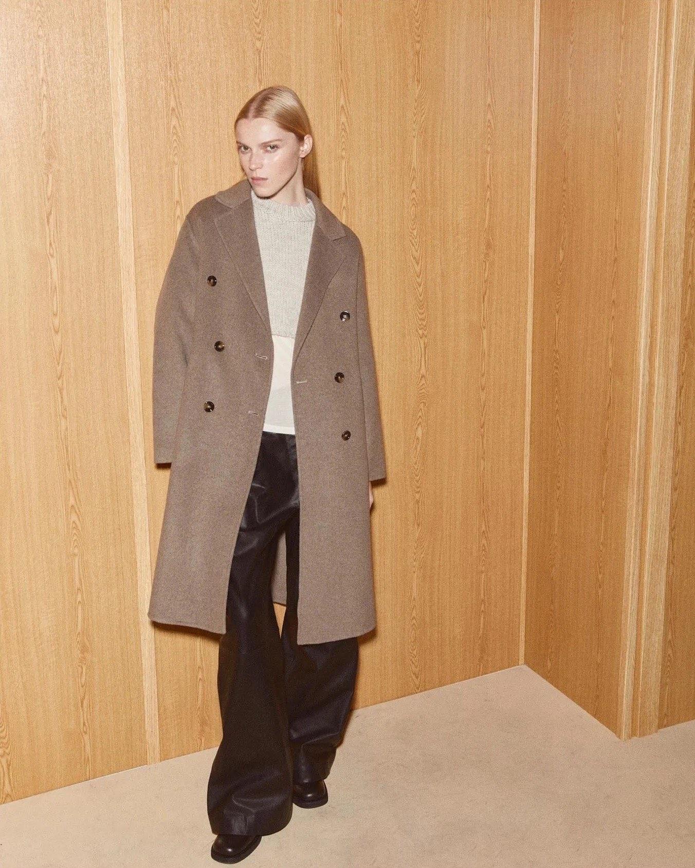 A model in a taupe double-breasted coat against pale wood paneling