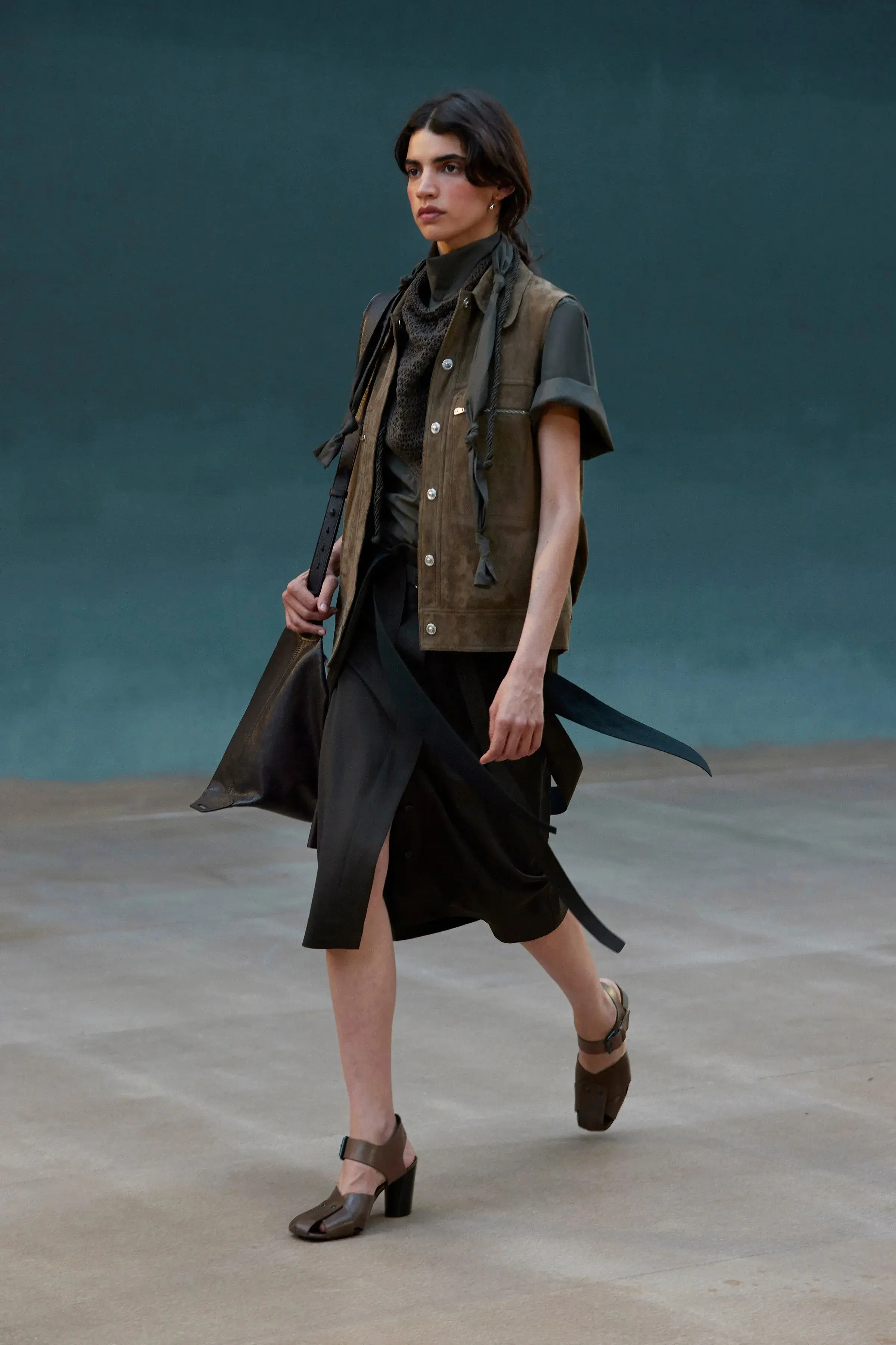 A model walking in an olive suede gilet over a wrapped skirt, carrying a soft leather bag