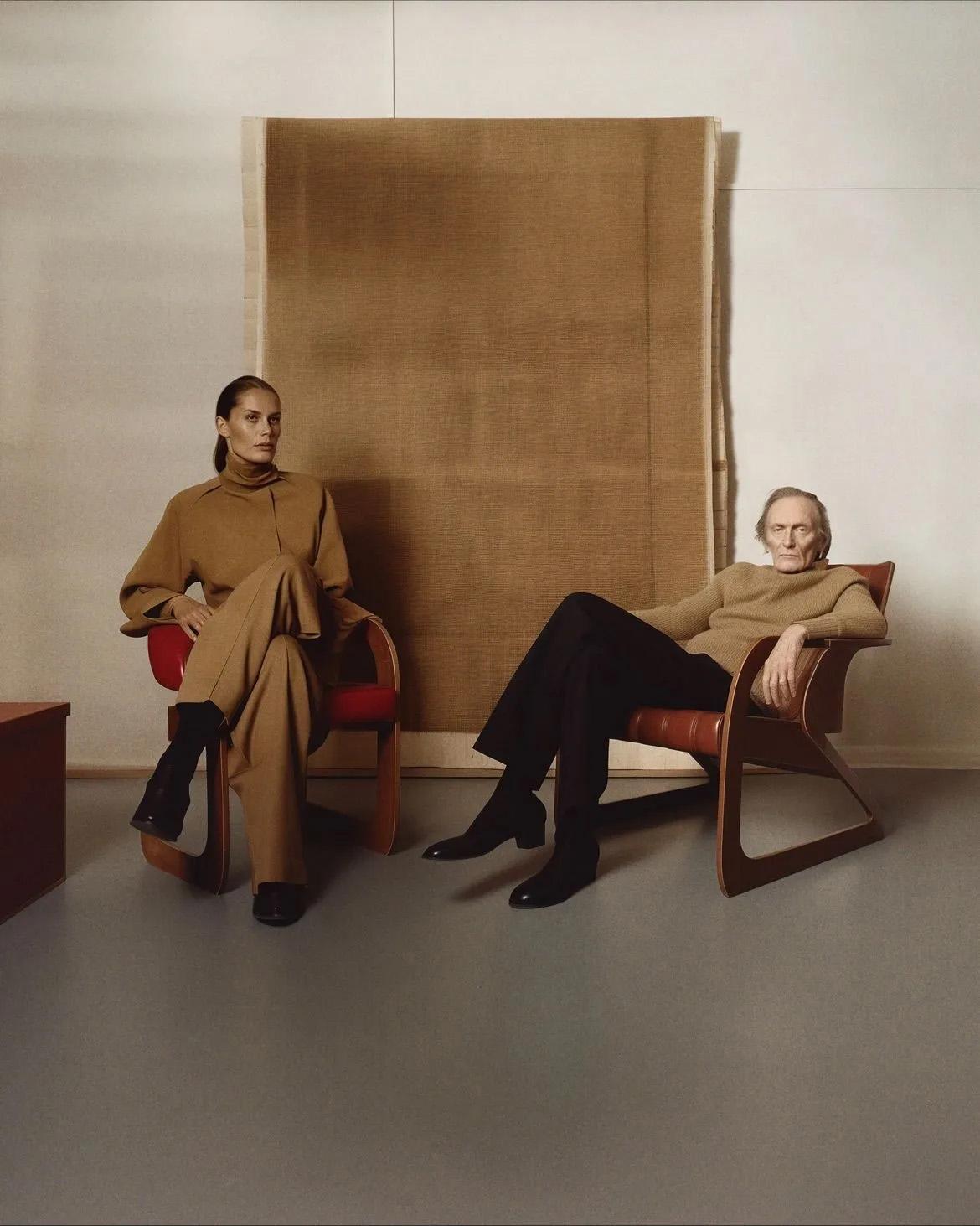 Two people seated in camel knitwear in front of a hanging woven panel